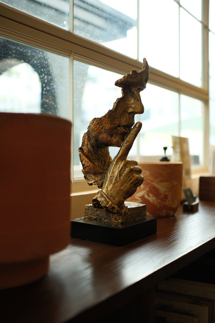 A rustic sculpture of a face and hand in a thoughtful pose by a rain-dappled window.